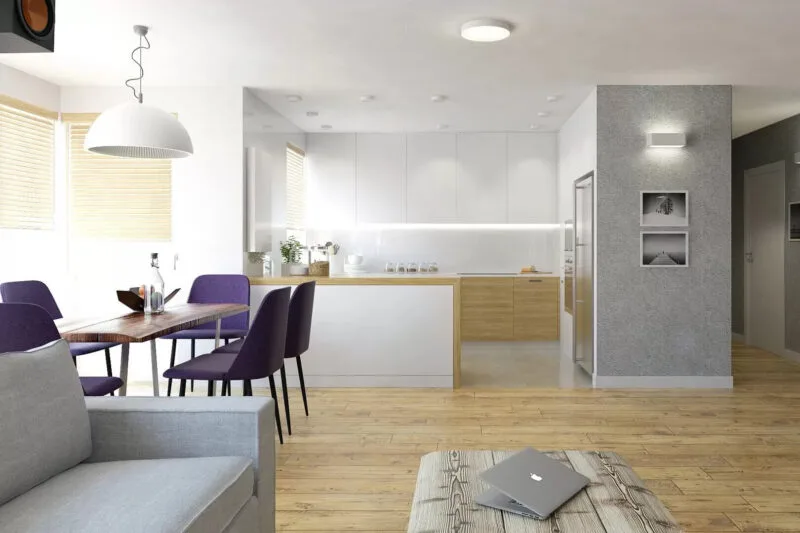 Jasny salon z aneksem kuchennym. Podłoga z jasnego drewna, szarą sofa, drewniany stół z pięcioma fioletowymi krzesłami. Po lewej stronie dwa okna z drewnianymi roletami. Na środku drewniany stół. Widać aneks kuchenny z szafkami w kolorach białym i brązowym. Na ścianie widać dwa obrazy. I po prawej stronie widać część korytarza i drzwi do następnego pokoju.