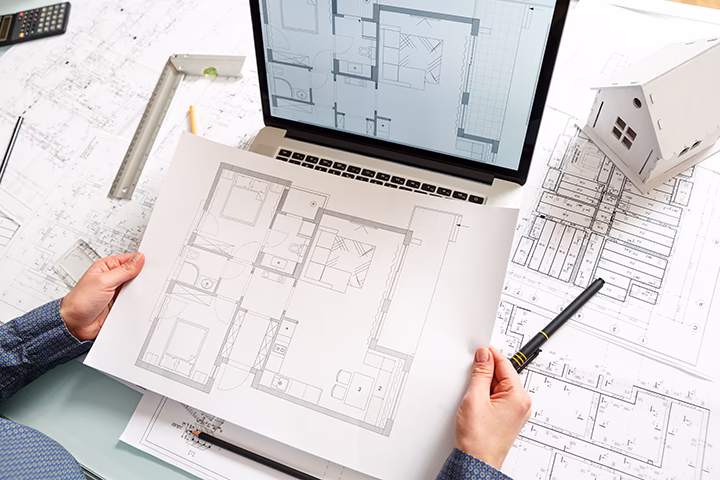 Architekt trzymający plan mieszkania nad laptopem i makietą domu.