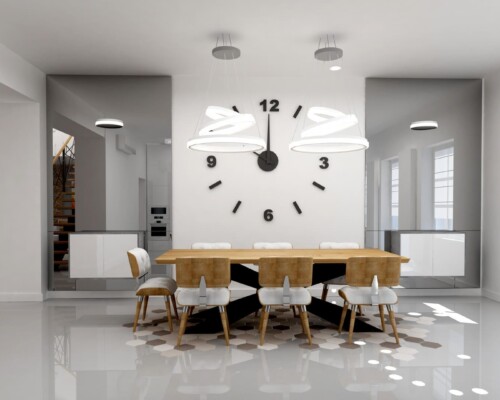Płynne przejścia i spójna estetyka – harmonia między strefą jadalni a otwartą kuchnią. Widok na nowoczesną jadalnię z zegarem i stołem, w tle widoczna biała kuchnia z wyspą.