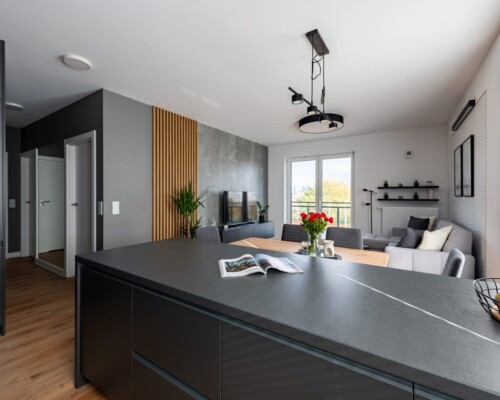 Kuchnia z wyspą – przestrzeń i styl w jednym Wyspa kuchenna z czarnym blatem, drewnianą wnęką i nowoczesnym oświetleniem