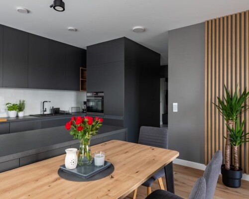 Kontrast i funkcjonalność – kuchnia z charakterem Ciemna kuchnia z wyspą, drewnianym stołem i nowoczesnym oświetleniem